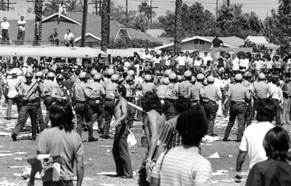 Phalanx of helmeted police with batons confront group of students in front of houses and palm trees.