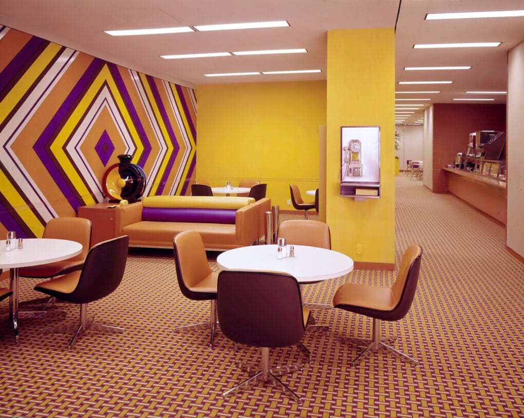 Carpeted cafeteria seating area with yellow wall and supergraphic wall in yellow, white, orange, and purple