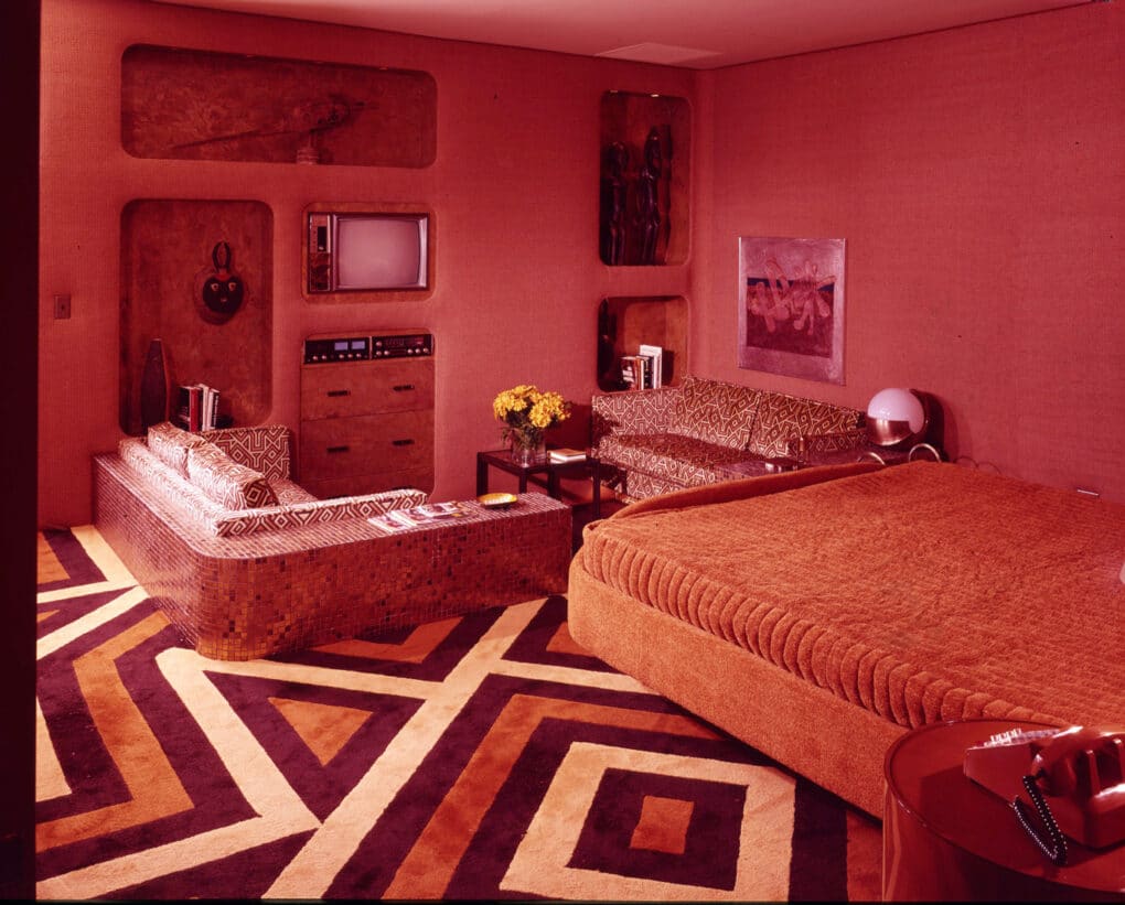Bedroom carpeted in red, brown, and white shag, with red bedspread, red walls, and built in sofa and television.