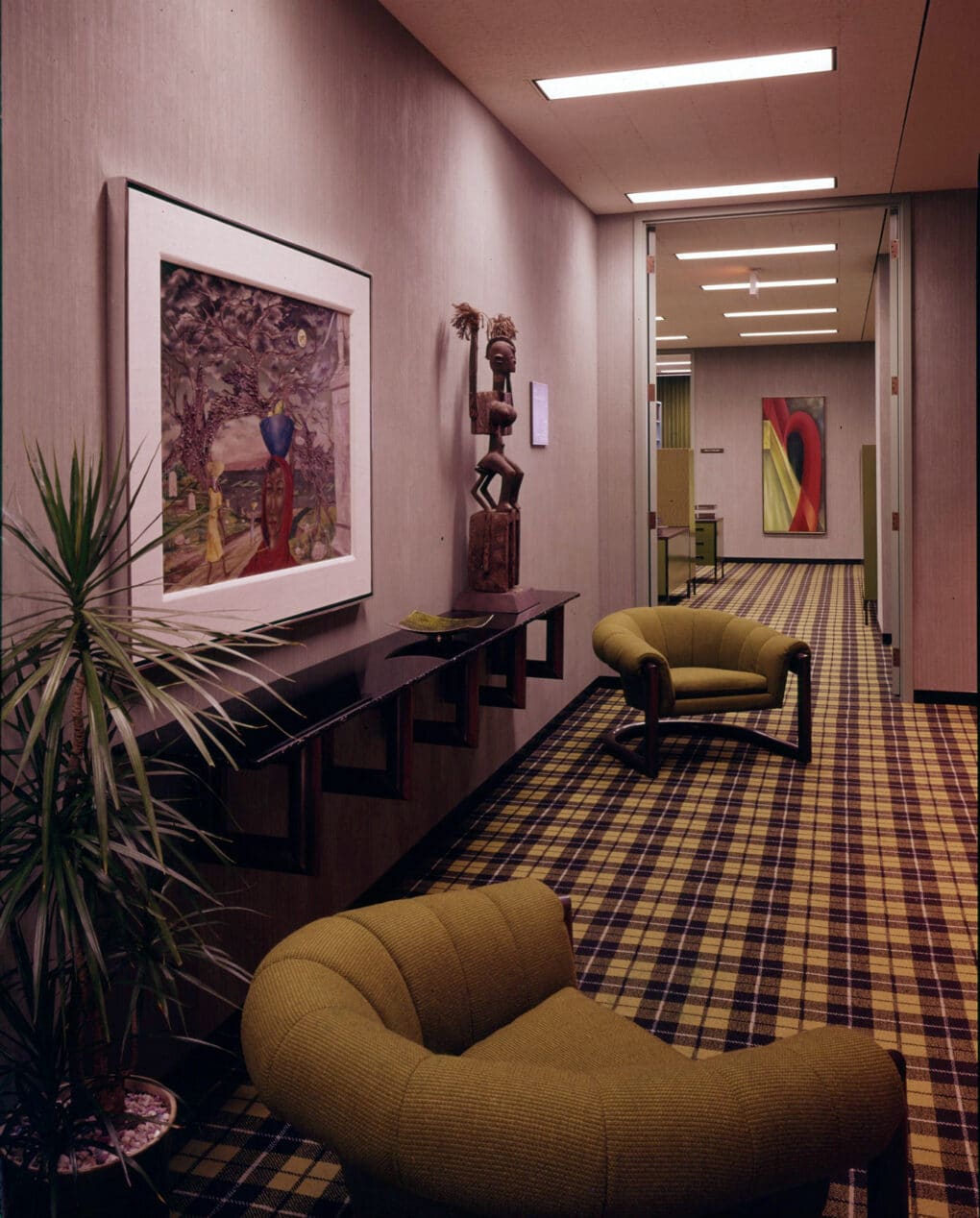 Office hallway with yellow and purple plaid carpet, yellow chairs, framed paintings, and African sculpture