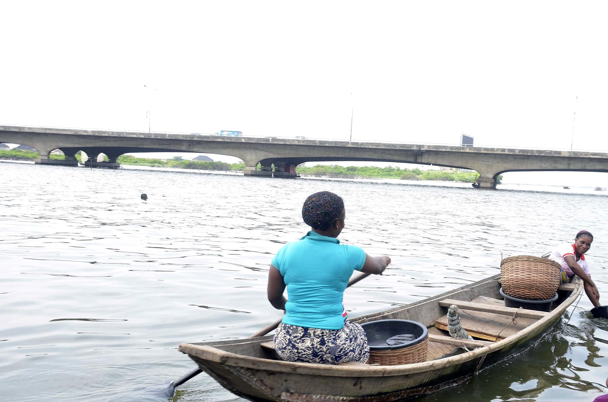 Click to Enlarge A woman paddles her canoe toward a bridge over a body of water.