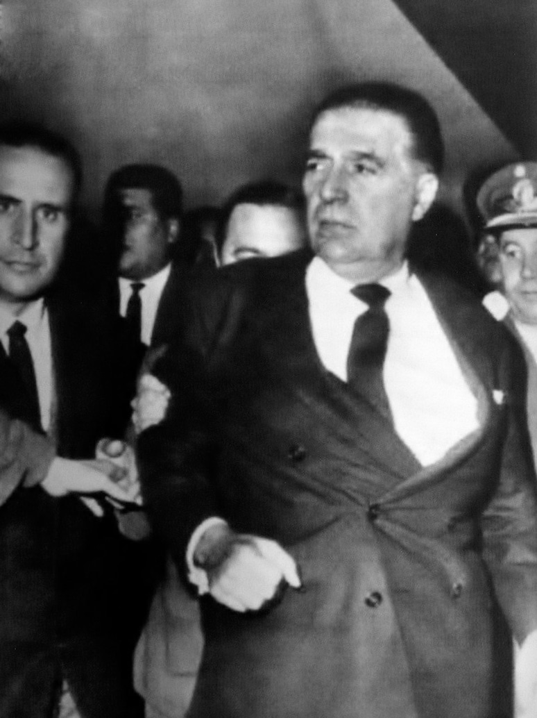 Black-and-white photograph of middle-aged white man in a suit being conducted by the arms by other men, one wearing a military uniform.