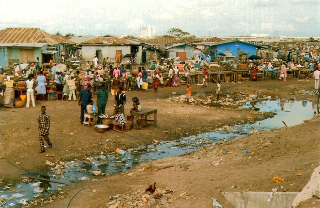 Concrete houses with tables set up in front of them; people browsing. In the foreground, a ditch running through a dirt lot.