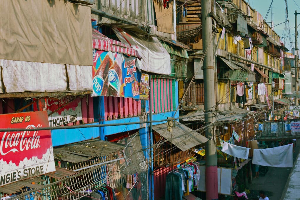 Densely packed street in informal settlement with advertisements, clothing, wires, and awnings hanging from balconies.