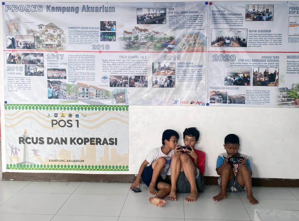 Three little boys looking at cell phones, sitting on a tile floor below a frieze-like poster with dates and photographs.