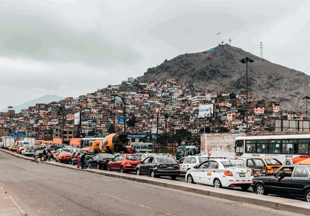 Triangular hill densely built halfway up with small colorful houses; in the foreground, bumper-to-bumper traffic on a highway.