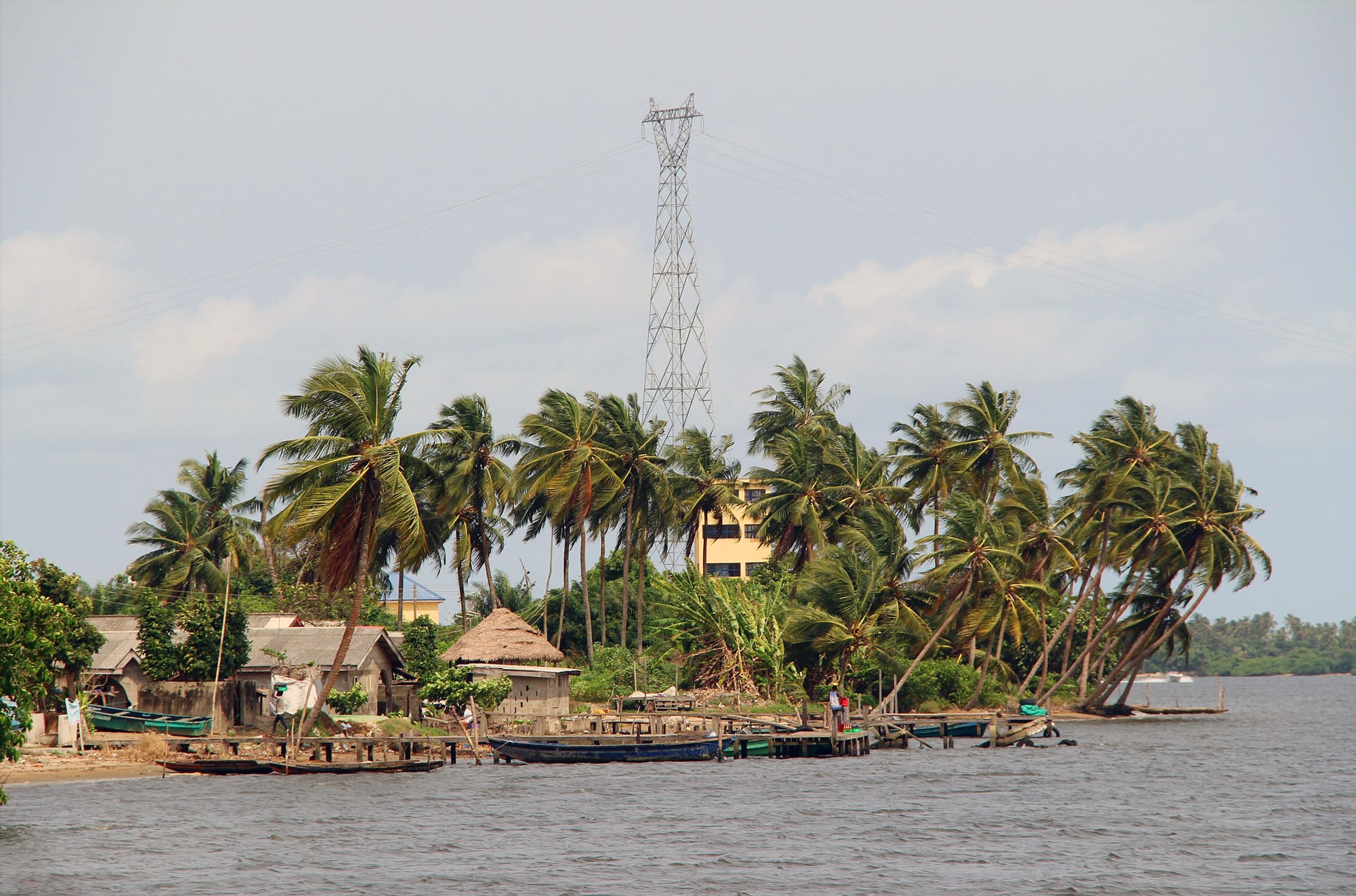 Click to Enlarge Huts and canoes sit by the water, beneath palm trees that are leaning with the wind. In the background, a cell tower.