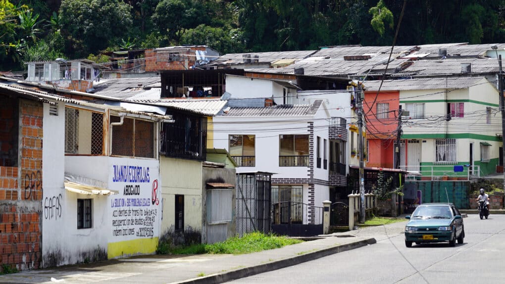 Street in a modest, densely built neighborhood, with a car and a person on a motorbike.