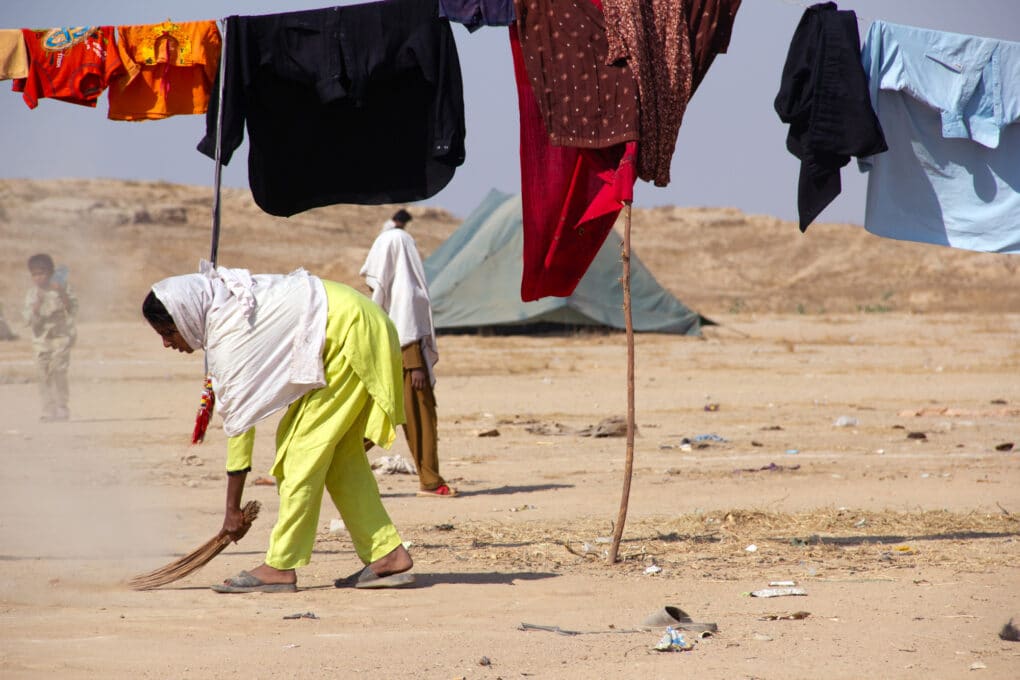 A woman sweeping dusty ground, with a clothesline over her head and in the back ground several other people and a tent.