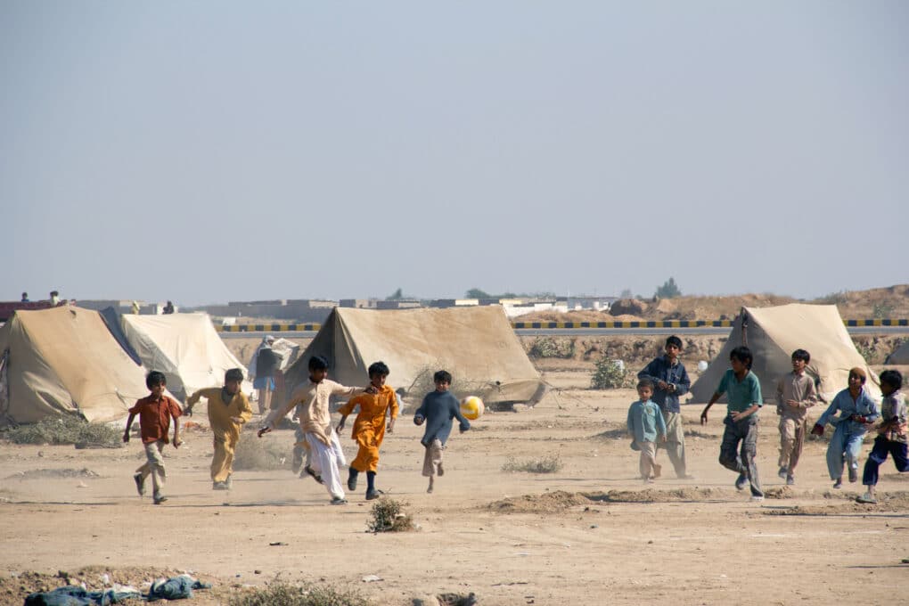 Children playing soccer in a dusty open space, with tents in the background.