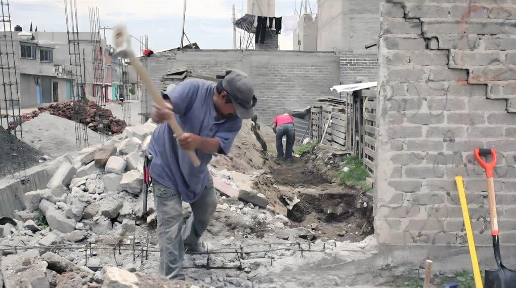 Two men demolishing a cinderblock wall in a densely built neighborhood.