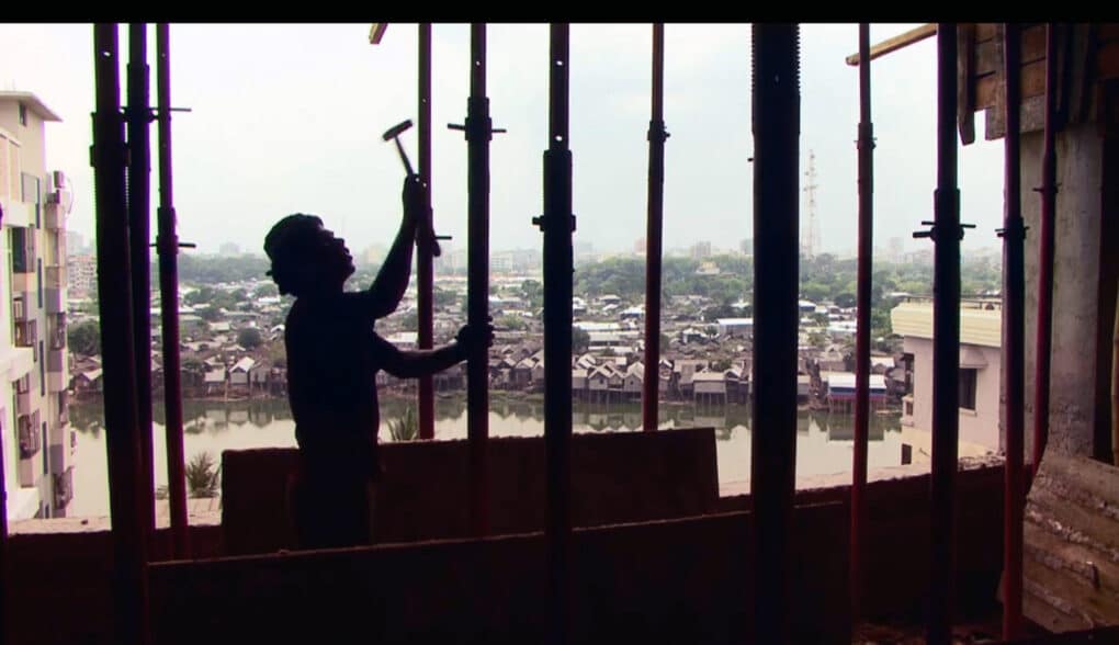 A man in silhouette erecting metal studs on a construction site; through the silhouetted bars a waterway and beyond that a densely crowded neighborhood of small shacks is visible.