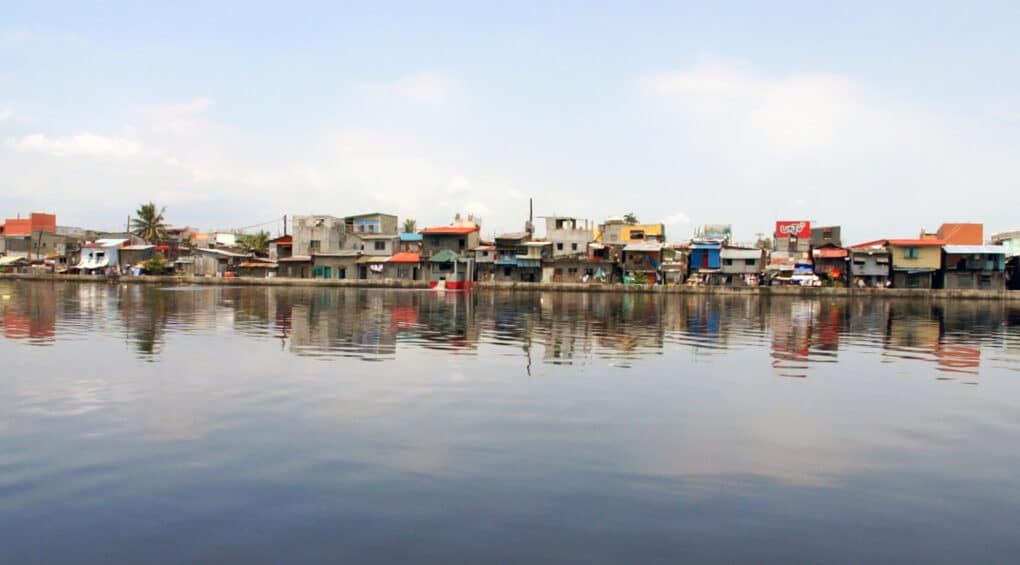 Densely built neighborhood of shacks photographed from the far side of a body of water that fills the foreground.