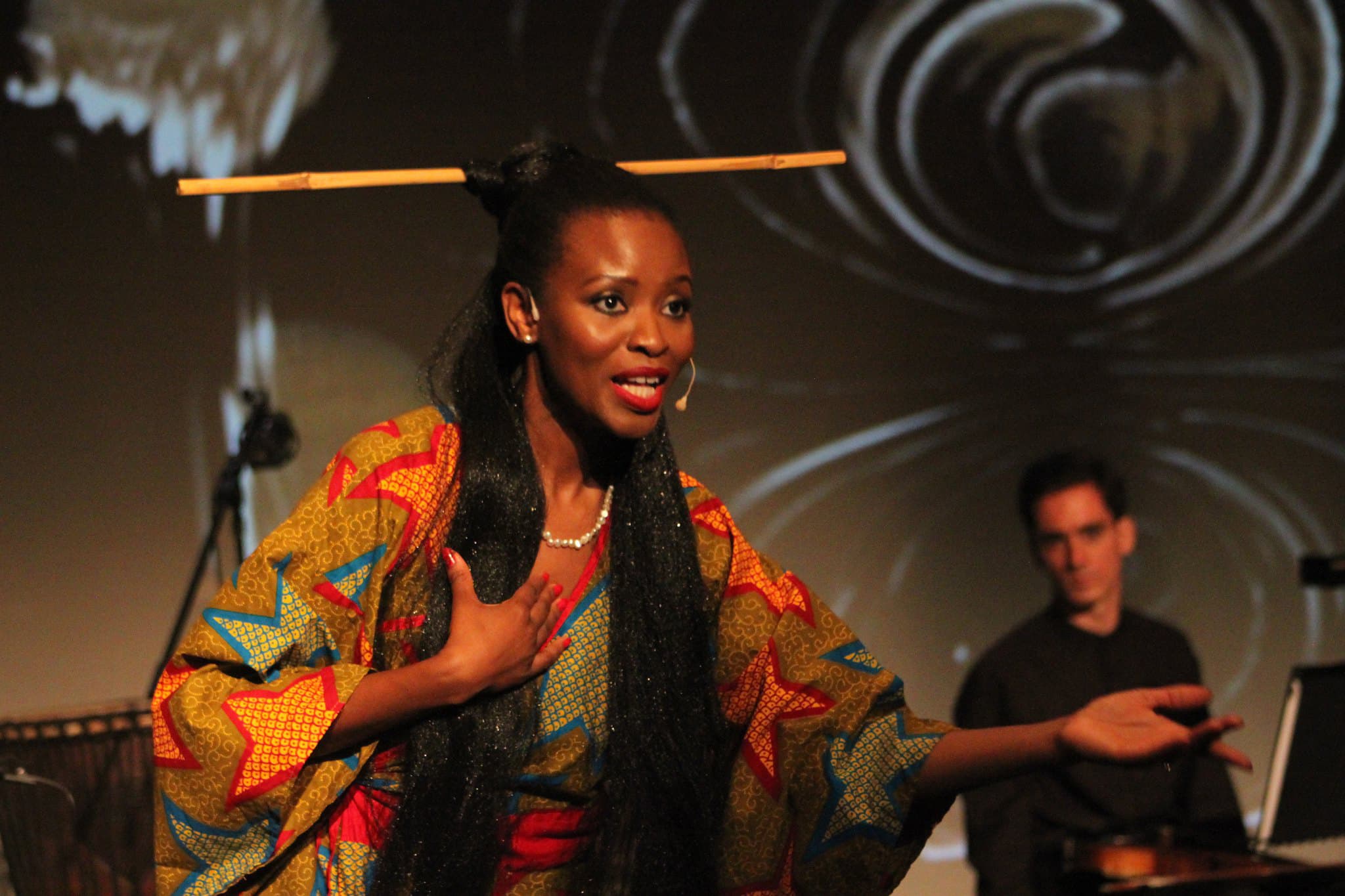 Click to Enlarge Black woman in printed gown performing onstage; musician in the background.