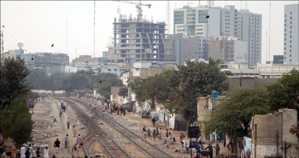 People walking along train line extending from the foreground toward the horizon, while behind it rise multiple concrete towers, one with a crane on top.