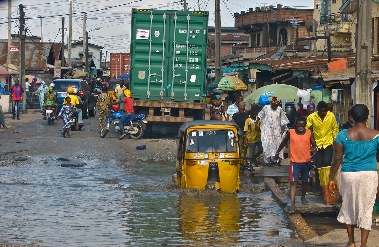 Click to Enlarge People balance on raised platforms as they traverse a flooded city street. A man sits in a car that is driving through a large puddle.
