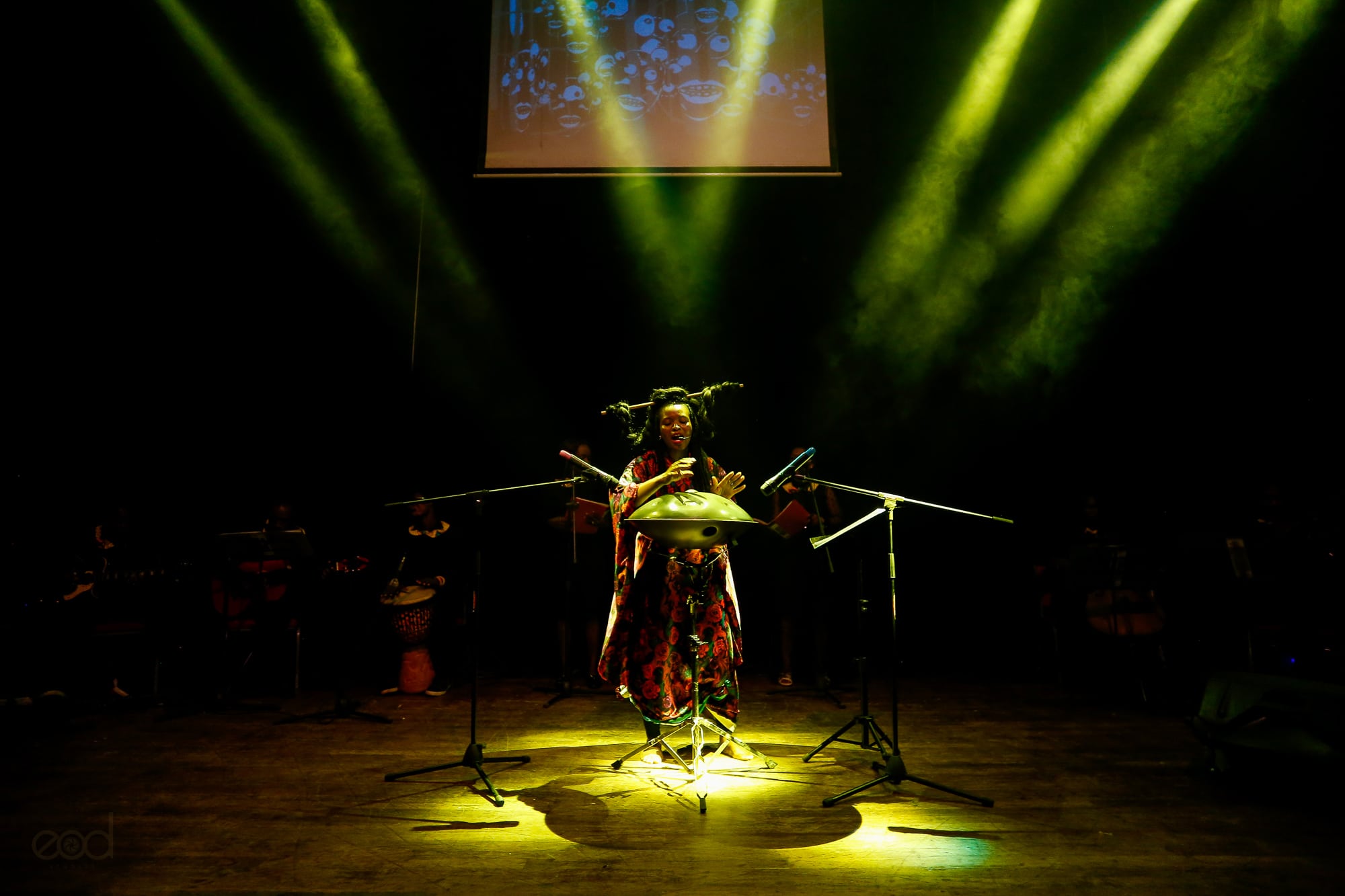 Click to Enlarge Black performer at a drum onstage, under dramatic green spotlights.
