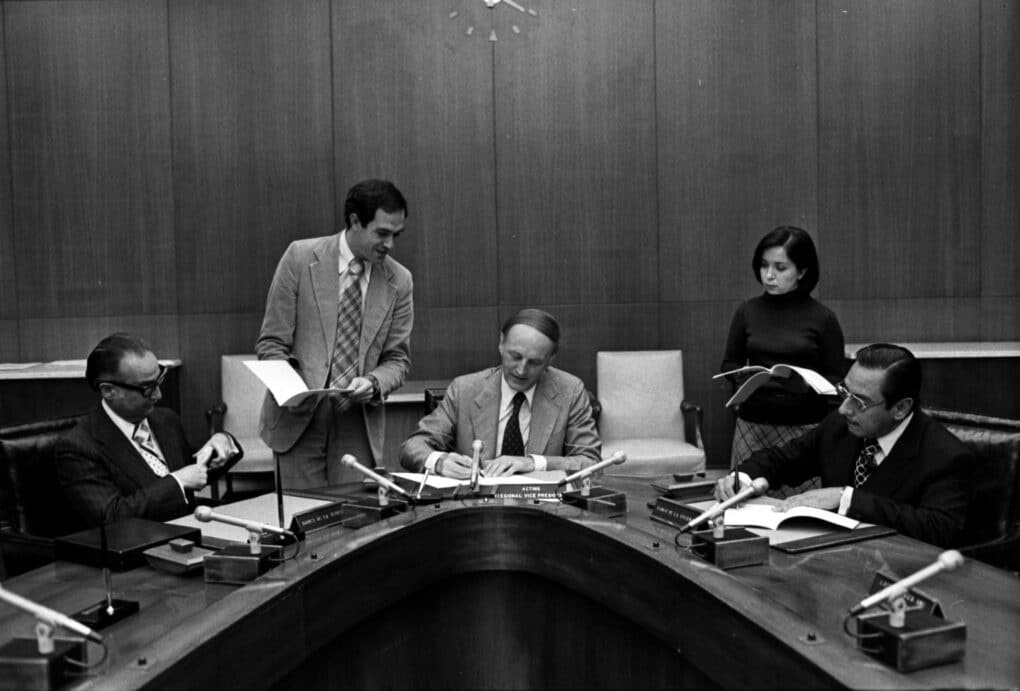 Black-and-white photograph of three men seated at a conference table, with a man and a woman standing behind them.