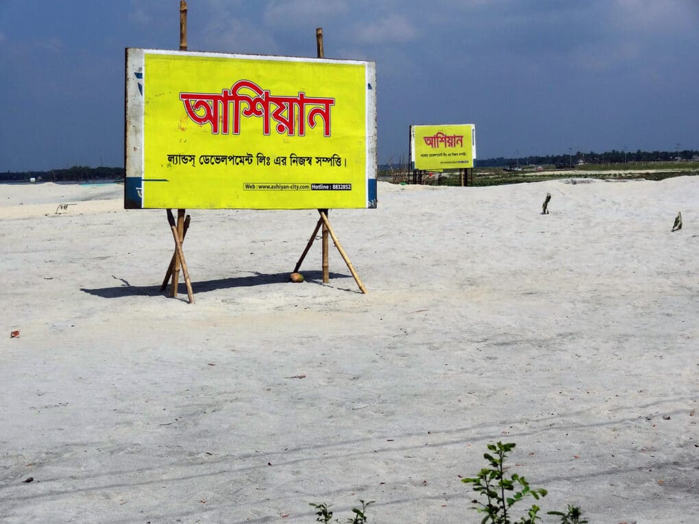 Color photograph of two yellow billboards with red and black writing in Bangla, standing alone in an empty sandy space.