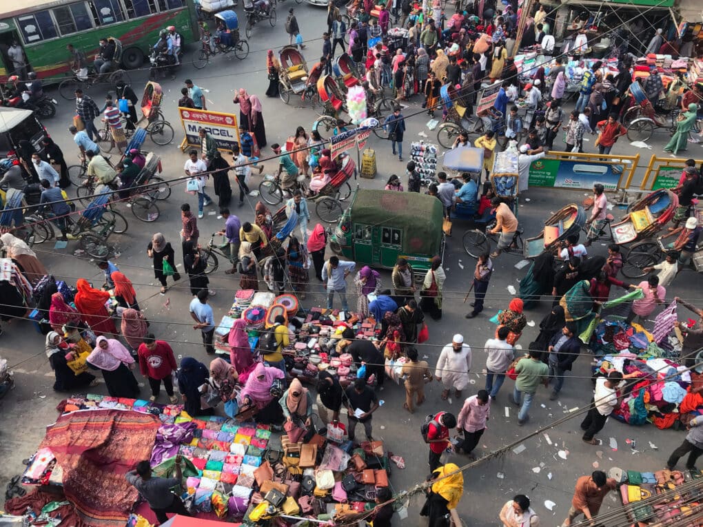 Overhead photograph of large crowd in an outdoor market, shopping at tables covered with brightly colored clothing and fabrics.