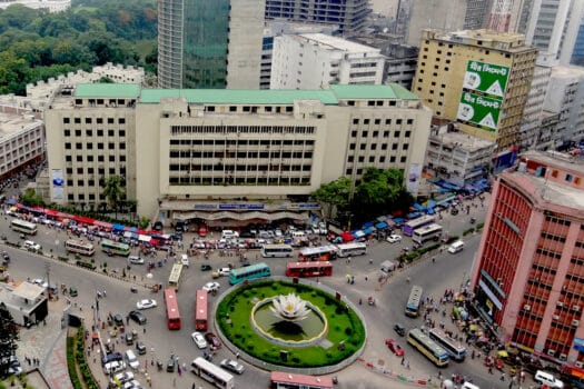 Color photo from an elevated perspective showing large roundabout in the center of a dense stand of skyscrapers. At the center of the roundabout stands a large sculpture of a lotus flower in a fountain pool.