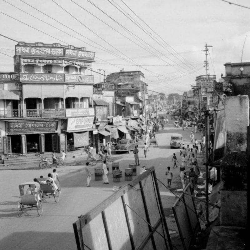 Click to Enlarge Black-and-white photograph of city street with low wooden buildings, some pedestrians, and traffic of bicycyle rickshaws.