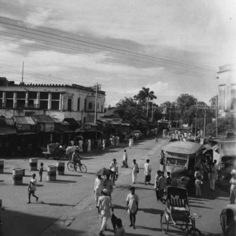Click to Enlarge Black-and-white photograph of city street with low wooden buildings, some pedestrians, and traffic of bicycyle rickshaws