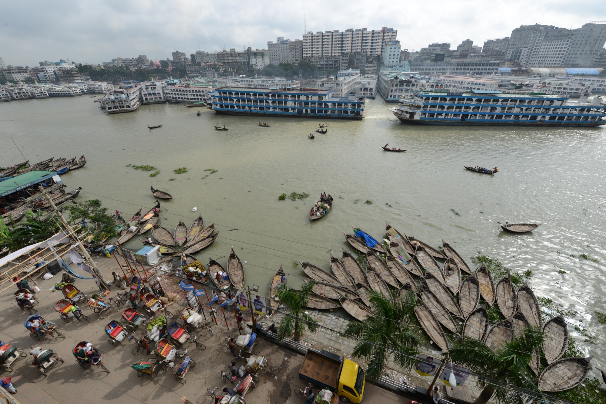 Click to Enlarge Color photograph of wooden boats and bicycle rickshaws at a riverside dock, with scattered wooden boats on the river and densely built city on the opposite shore.