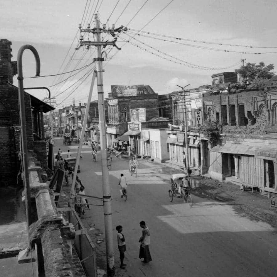 Click to Enlarge Black-and-white photograph of city street with low wooden buildings, some pedestrians, and traffic of bicycyle rickshaws