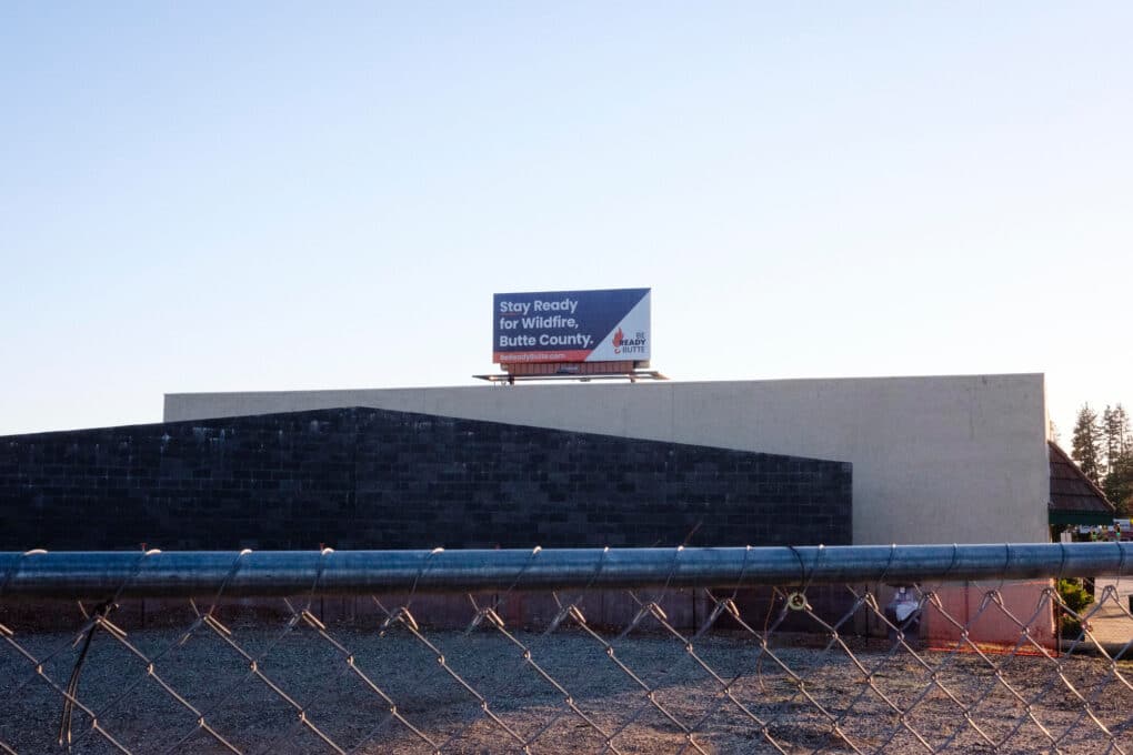 Color photograph of municipal building behind chainlink fence; a billboard on the roof of the building reads: "Stay Ready for Wildfire. Butte County."