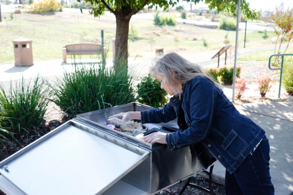 Color photograph of a middle-aged white woman in a jean jacket placing small items in an open metal box in a small park.