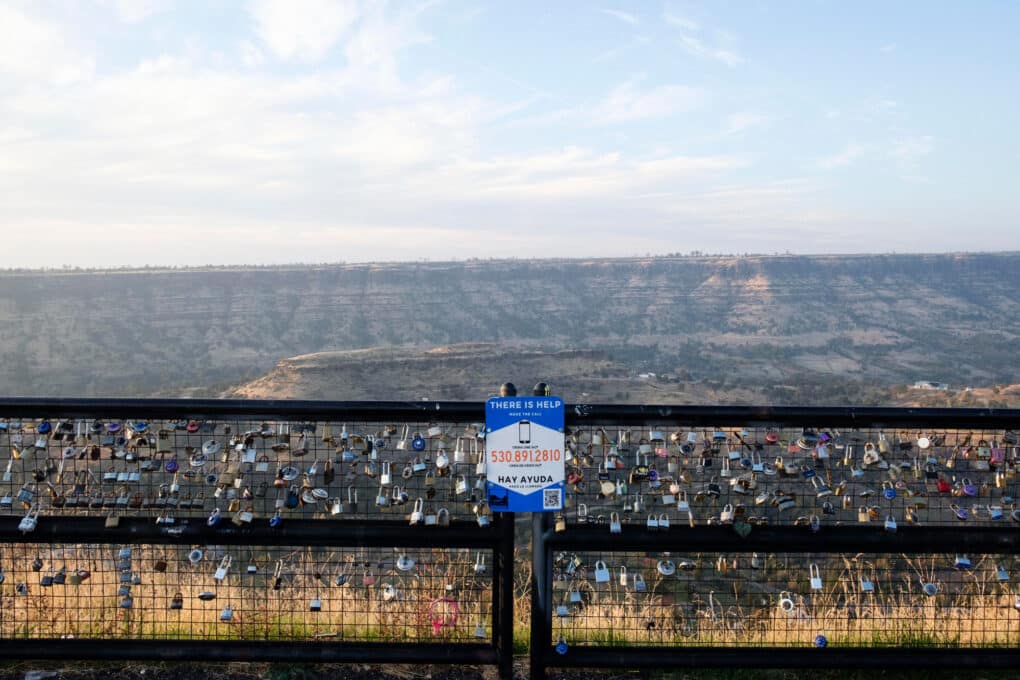 Color photo of dark, burned ravine, taken from an overlook point. The guardrail fence is thickly lined with padlocks. In the center is a sign reading "There is Help" with a phone number.