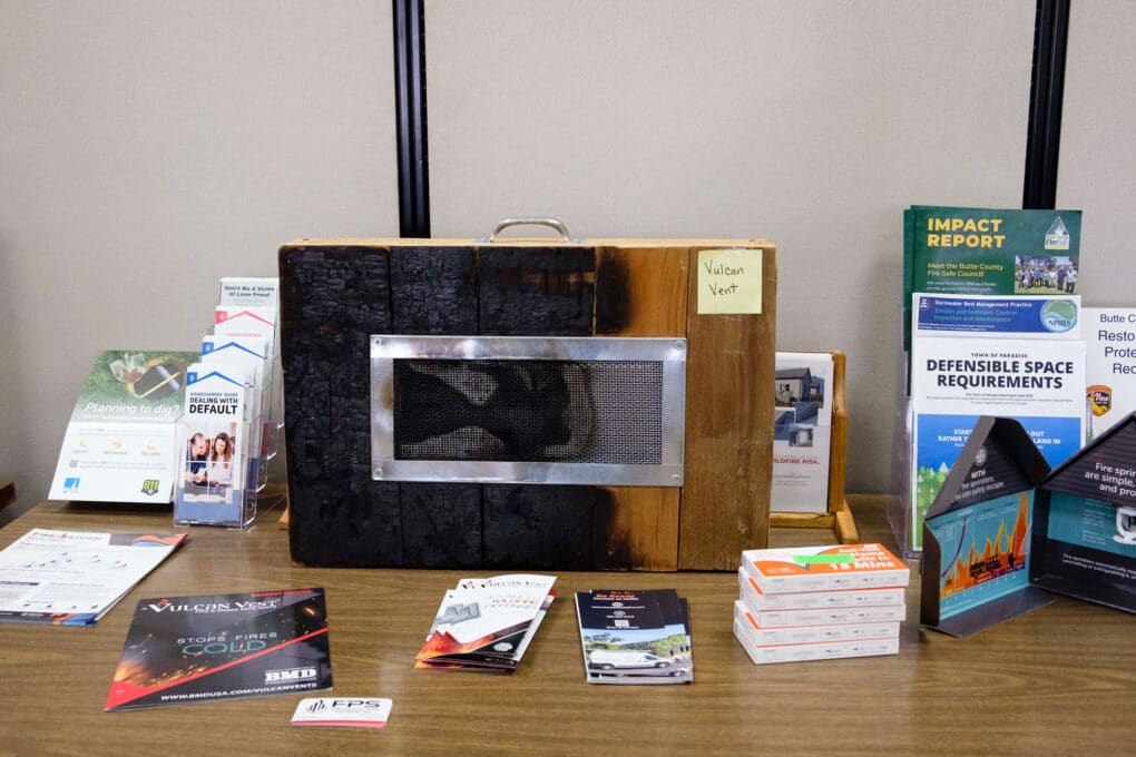 Color photograph of display in an office, including brochure titled "Defensible Space Requirements," a stack of covid test boxes, and a partly-burned display unit with a vent set into wooden siding, labeled "Vulcan Vent."