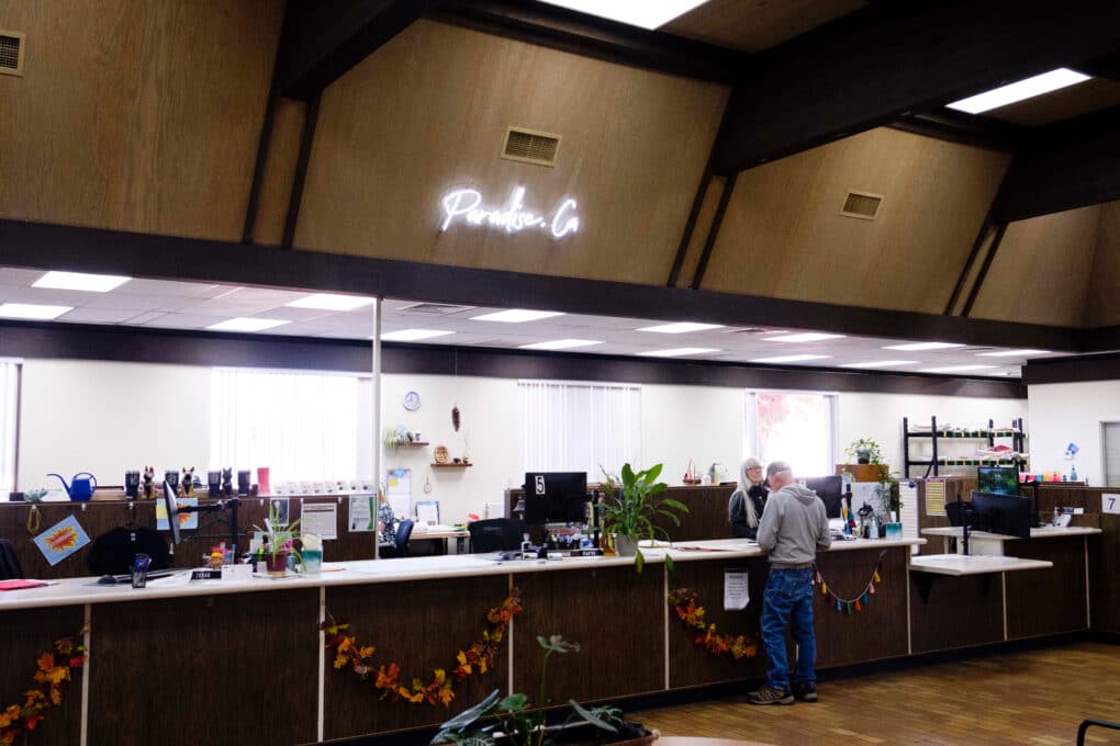 Interior of a municipal office, showing a man in hoodie and baseball cap being helped by a woman behind the counter. Over their heads, a white neon sign reads "Paradise CA."
