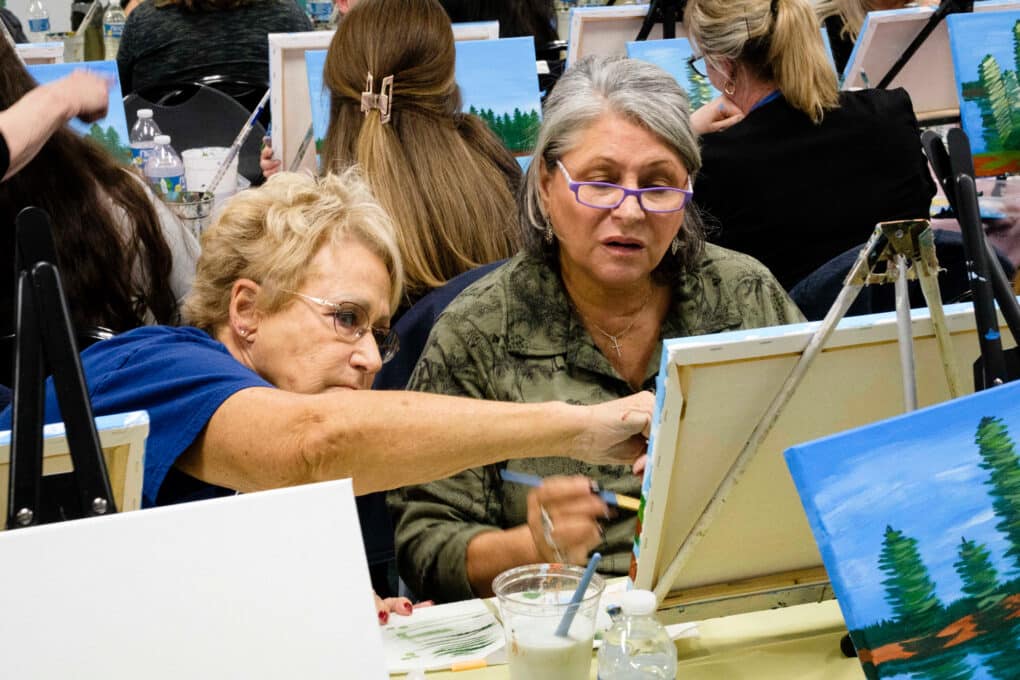 Color photo of a group of white women of mixed ages in a painting class--each painting shows blue sky and pine trees.