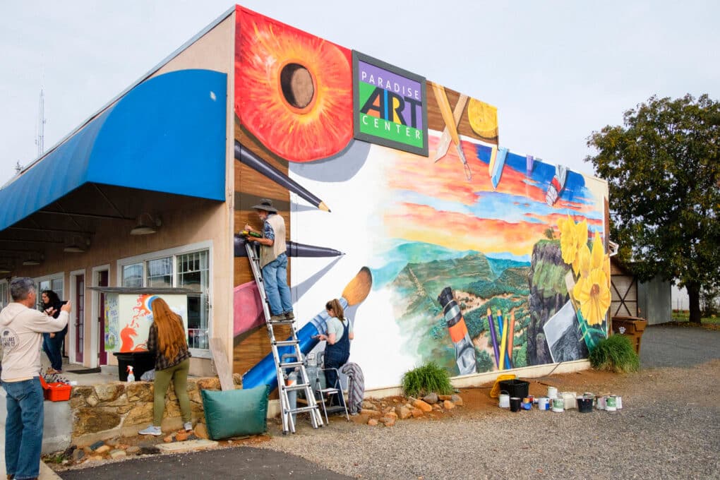 Color photograph of a small building with a sign reading Paradise Art Center. A small group of people are painting a mural on the building's side wall.