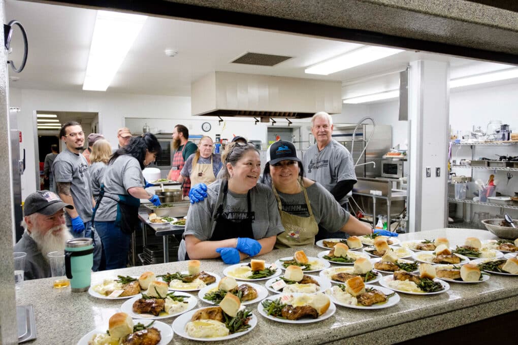 Color photograph of a group of white people of mixed ages at an industrial kitchen pass-through, with plates of food set up on the counter.