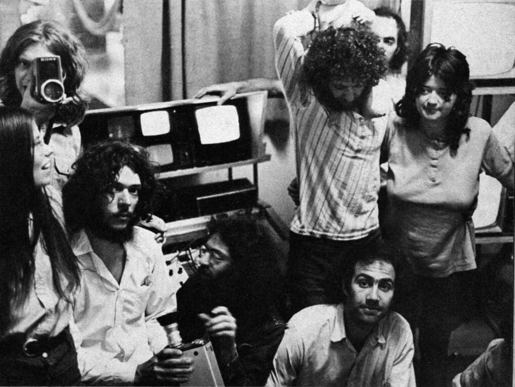 Archival 1970s black and white photograph. Young people gathered around video monitors. One person points a camera at the photographer.