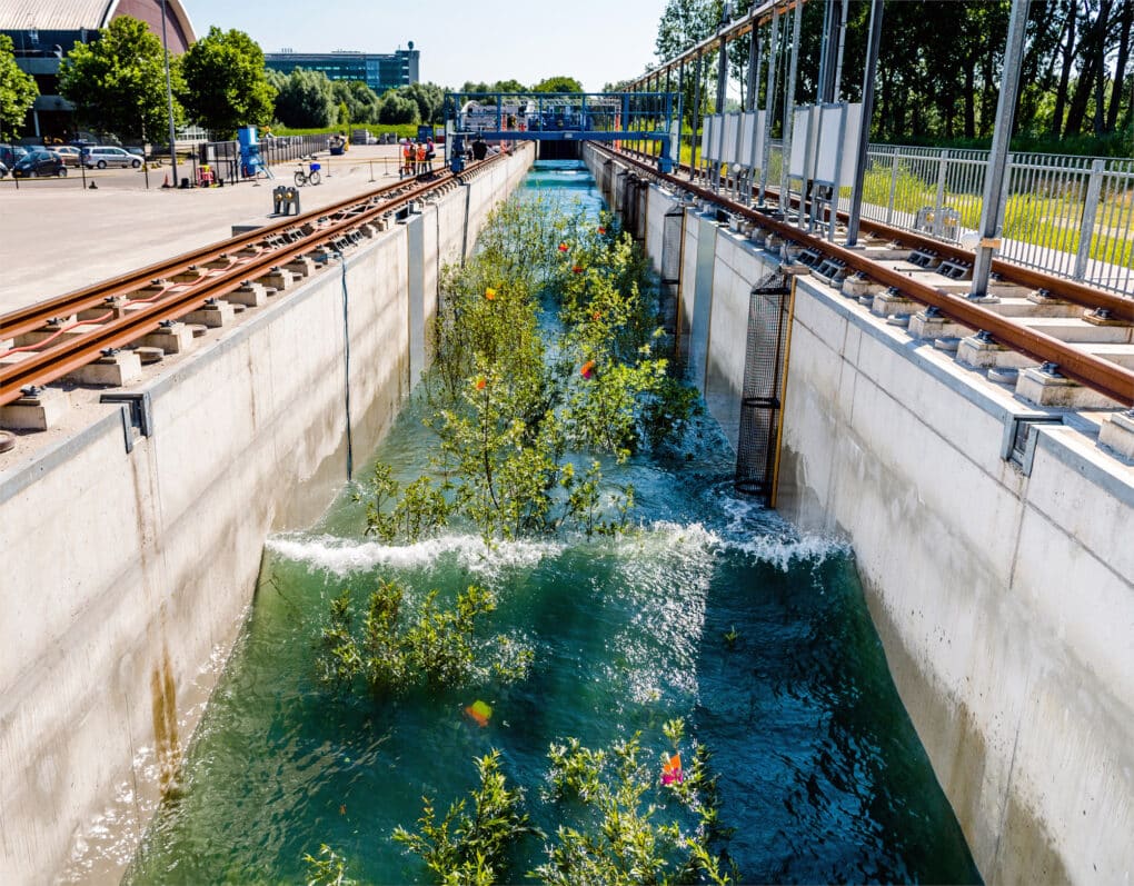 Outdoor hydraulic model. Long concrete channel with water. A wave is going down the channel, passing through small trees.