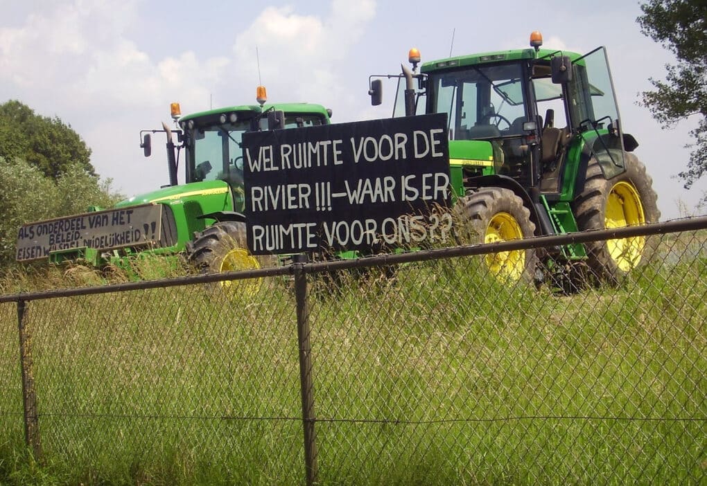 Two tractors parked behind in tall grasses behind a chain-link fence. A protest sign reads, in Dutch, "THERE IS ROOM FOR THE RIVER!!! WHERE IS ROOM FOR US?"