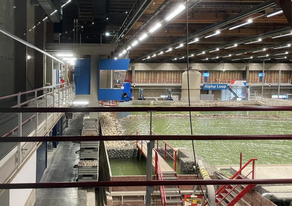 Hydraulic model in a large indoor space. From a catwalk, we see a large pool of green water with small waves, as well as a line of rocks that simulates a jetty.