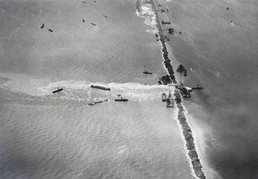 Black and white photo from an aerial perspective, with dozens of sea vessels working on the construction a long dam