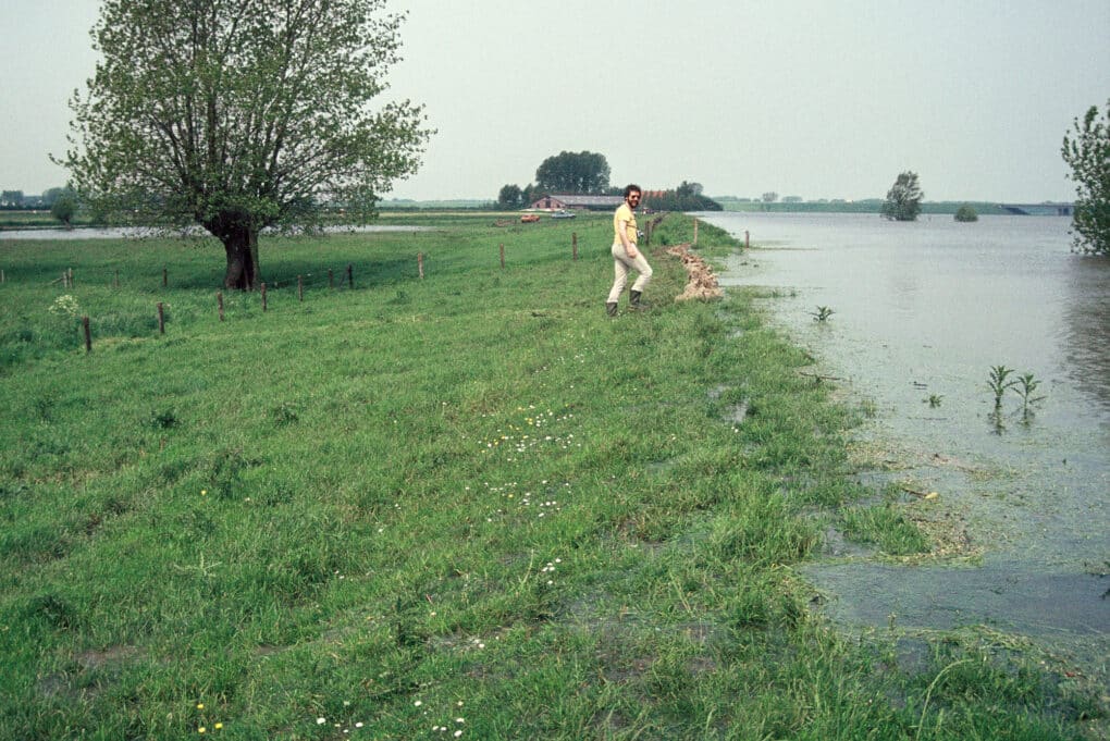 Man walking in a field next to a flooded canal that has just reached the top of an earthen dike.