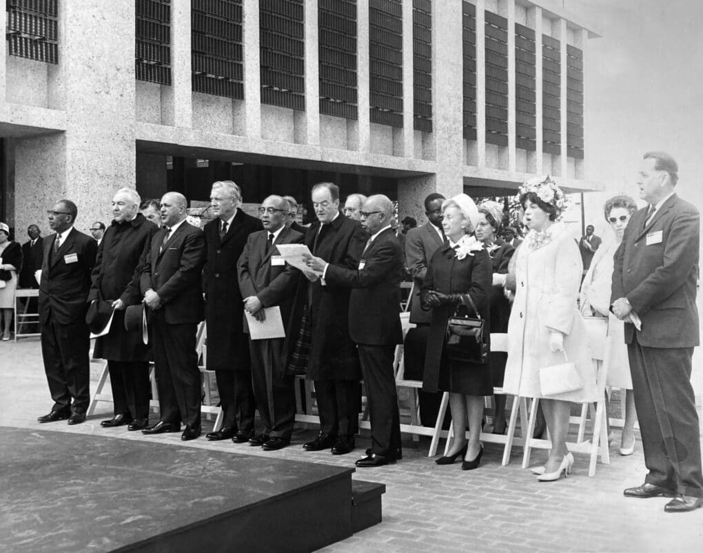 Black and white photo of a well-dressed group of people, black and white, standing in an outdoor audience in the courtyard of a modernist building. They appear to be singing.