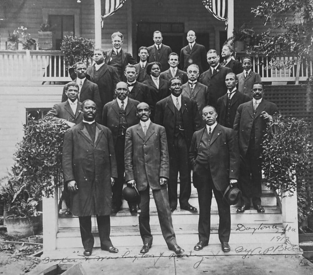 Group of black businessmen (plus one black woman and a few white men) standing on the steps of a white wooden building draped with American flags.