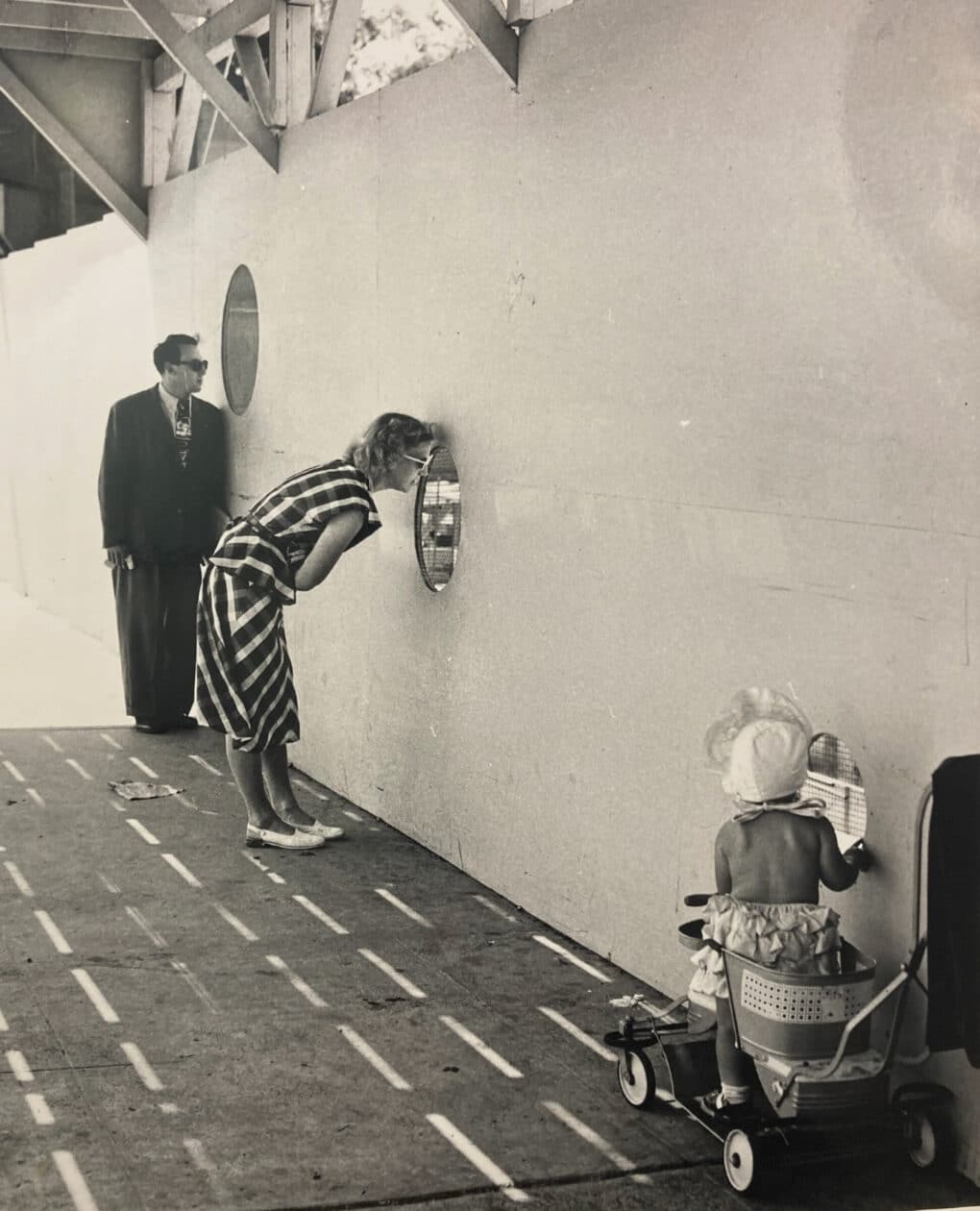 Black and white photo of white father, mother, and toddler in a stroller, each looking through a round opening in a construction hoarding.