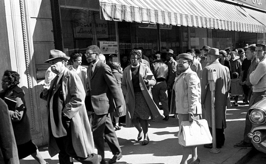 Black and white photo of well-dressed mixed-race crowd on the sidewalk outside a storefront, with a striped awning overhead.