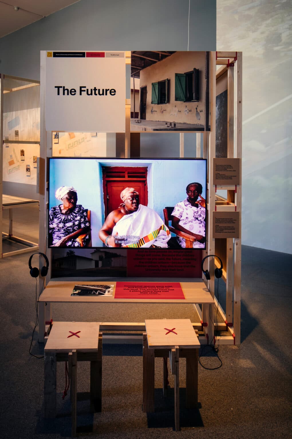 Color photograph of exhibition installation with video, showing three middle-aged African women seated in authoritative poses; above the screen, signage reads, "The Future."
