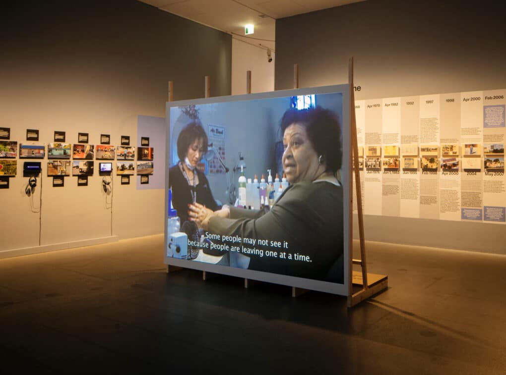 Color photograph of exhibition interior with large projected video, timeline, and small photos and videos on small wall-mounted monitors.