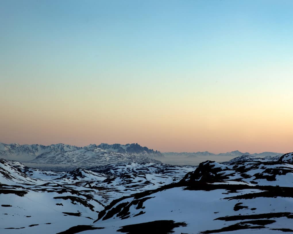 A color photograph of snowy, rocky horizon line at sunset with blue skies turning peach colored.